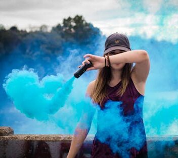 A stylish woman surrounded by bright blue smoke outdoors, creating a dynamic fashion statement.
